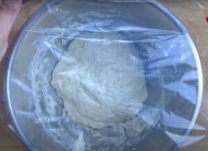 Bowl of dough covered with plastic wrap, ready to rise for homemade bread recipe on wooden surface.